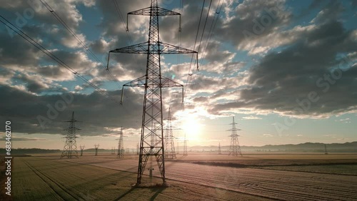 Electricity transmission towers with sunlit sky at sunrise aerial view. High voltage power pylon in field with nature landscape and cloudscape in morning drone shot photo