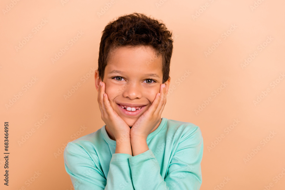 Photo of adorable charming boy wear trendy clothes arms touch cheeks face isolated on beige color background Photo of adorable charming boy wear trendy clothes arms touch cheeks face isolated on beige color background
