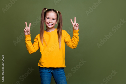 Portrait of satisfied pleasant cute small kid with tails dressed yellow sweatshirt showing v-sign isolated on khaki color background