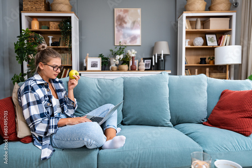 Young female freelancer in casual clothes sitting on comfortable couch and working remotely on laptop. Woman copyright writer or web developer work at home sitting on sofa enjoying her freedom.