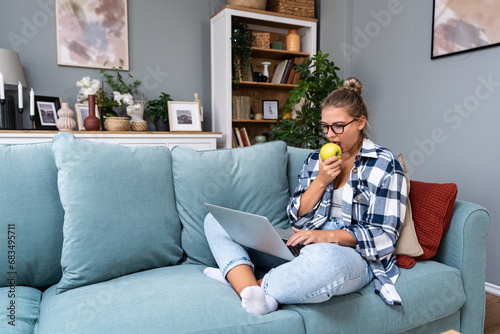 Young female freelancer in casual clothes sitting on comfortable couch and working remotely on laptop. Woman copyright writer or web developer work at home sitting on sofa enjoying her freedom.