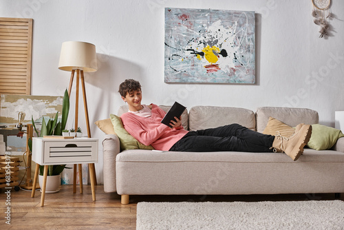 happy transgender person in pink sweater writing in notebook and sitting on sofa next to smartphone