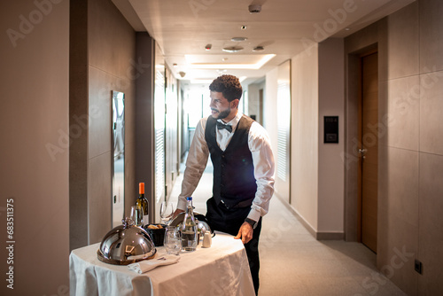Young waiter delivering tray with food in a room of hotel doing room service photo