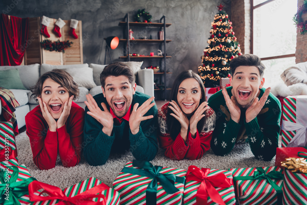 Fototapeta premium Full body portrait of group impressed positive fellows lying floor raise hands open mouth christmas giftbox decorated house indoors