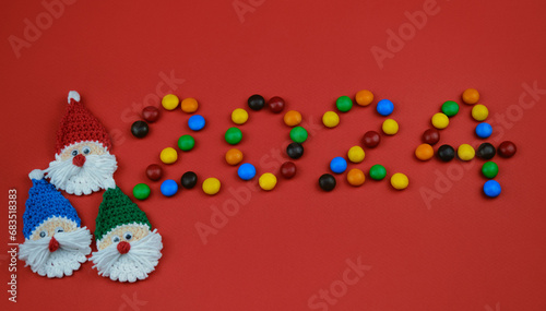 Happy New Year red background, inscription 2024 laid out of round chocolate colored candies. Minimalistic Christmas greeting card with copy space. Knitted toy amigurumi Santa Claus head.