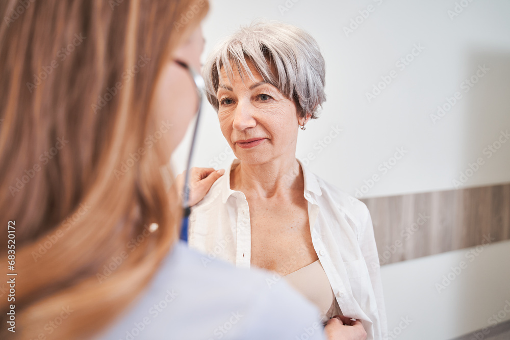Calm aged woman standing and professional female general practitioner using stethoscope Calm aged woman standing and professional female general practitioner using stethoscope