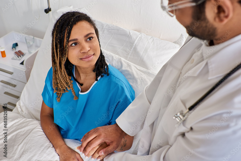 focus on jolly african american woman in hospital bed holding hand of her blurred indian doctor
