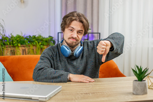 Dislike. Upset unhappy Caucasian man showing thumbs down sign fail gesture, expressing discontent, disapproval dissatisfied bad work at home office indoors. Displeased young guy in room sits at table photo