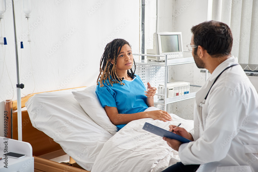 Fototapeta premium attractive ill african american woman lying in hospital bed talking to her young indian doctor
