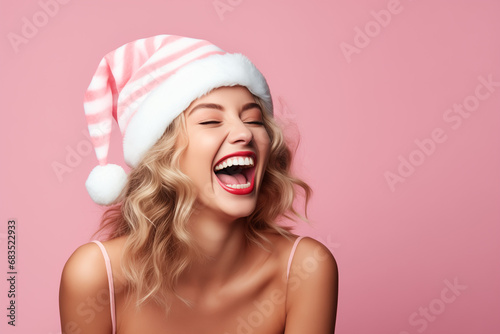A beautiful woman wearing a Santa hat and laughing on a pink background.