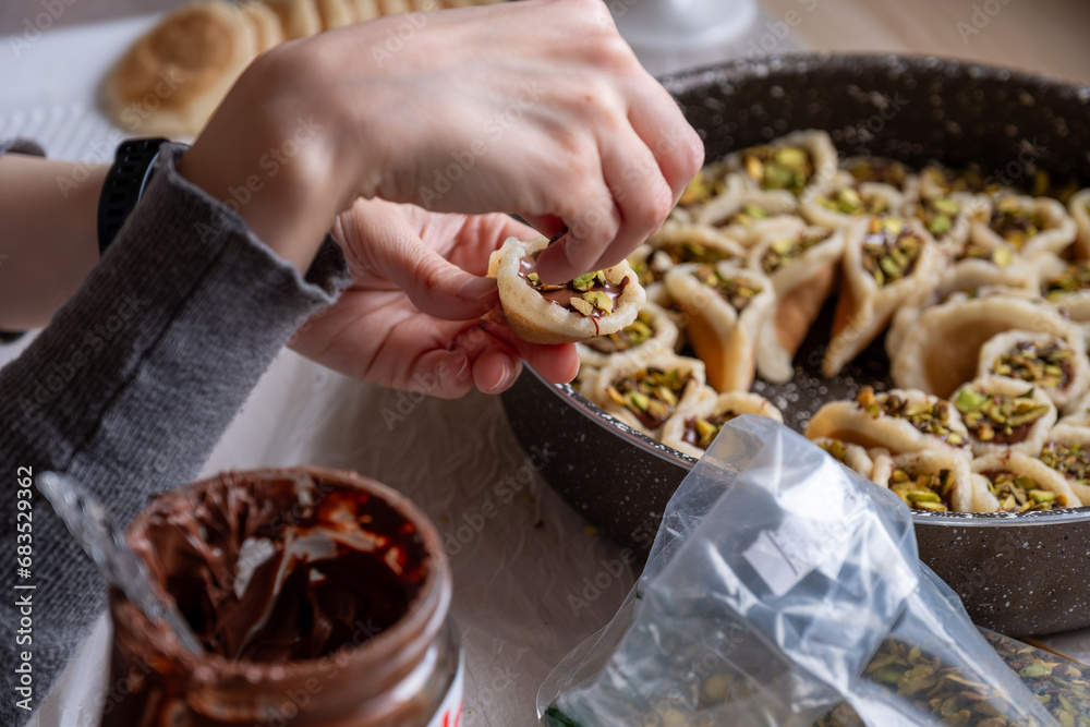Hands holding Qatayef filled with chocolate and topped with pistachios on a wooden table, with a plate to be prepared in the oven later as Ramadan sweets.