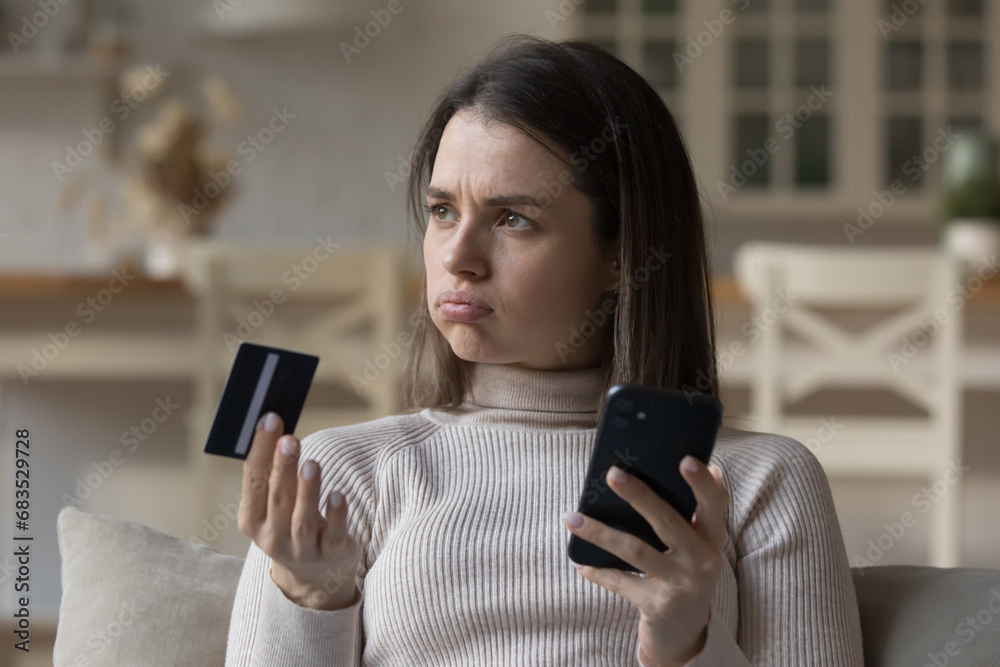 Frustrated young woman sit on sofa hold credit card use phone try to pay through e-bank app, experiences problem with e-payment due insufficient funds, no money, scam, unsuccessful transaction concept - obrazy, fototapety, plakaty 