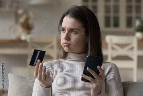 Frustrated young woman sit on sofa hold credit card use phone try to pay through e-bank app, experiences problem with e-payment due insufficient funds, no money, scam, unsuccessful transaction concept