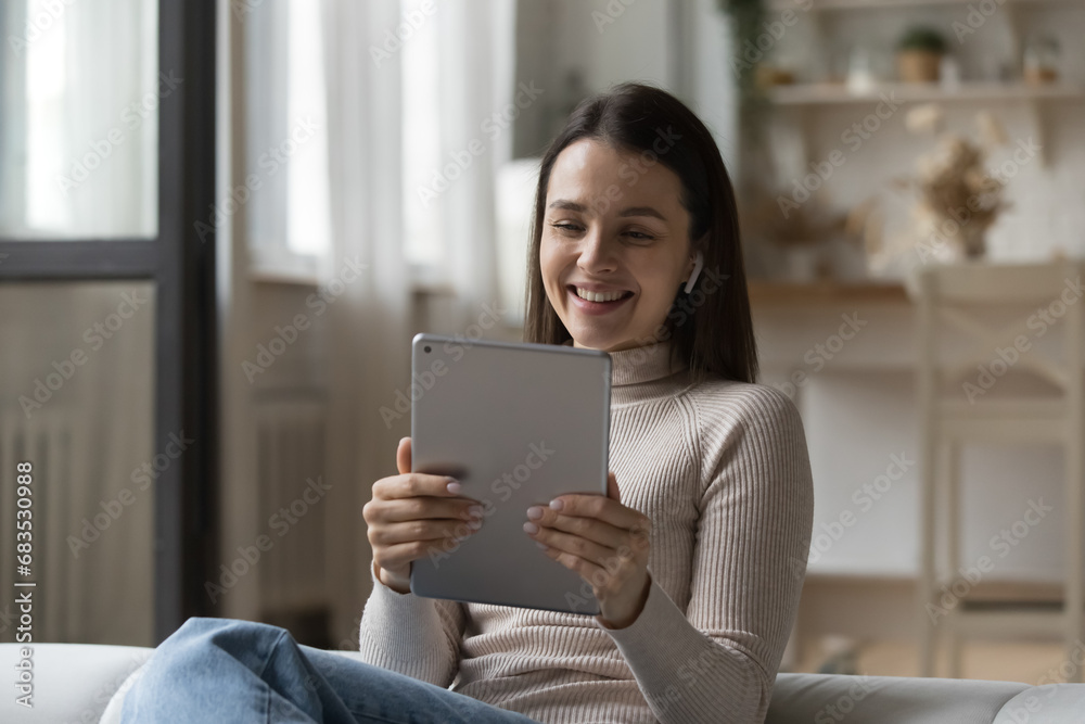 Young woman sit on sofa wear wireless earphones looks at digital tablet screen smile watch funny videos spend weekend leisure at home enjoy pleasant conversation use video call application on device. Young woman sit on sofa wear wireless earphones looks at digital tablet screen smile watch funny videos spend weekend leisure at home enjoy pleasant conversation use video call application on device.