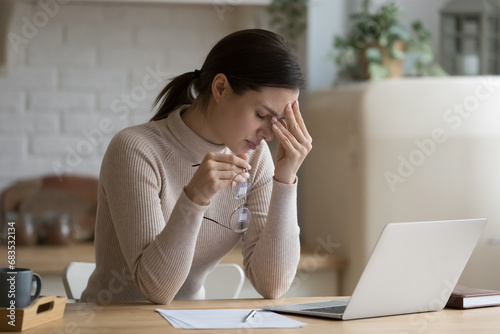 Young woman work on laptop feels overworked takes off glasses relieving eye strain massages nose bridge suffers from headache due to long usage of device. Dry itchy eyes need drops, eyestrain concept photo