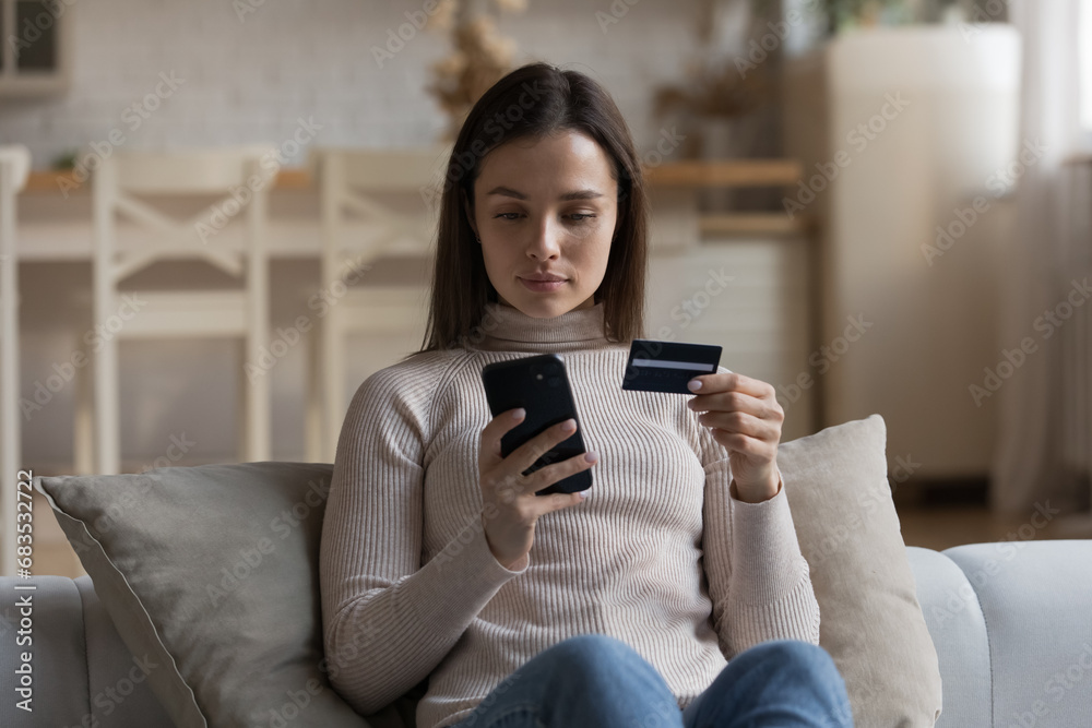 Serious woman sit on sofa holding cellphone and credit card buy goods make order on internet use retail e-services. Secure payment through e-bank app, instant money transfer, e-commerce client concept Serious woman sit on sofa holding cellphone and credit card buy goods make order on internet use retail e-services. Secure payment through e-bank app, instant money transfer, e-commerce client concept