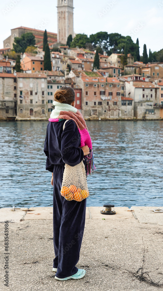 Girl in navy blue velvet costume holding bright orange tangerines in a beige string bag with beautiful mediterranean city on background .