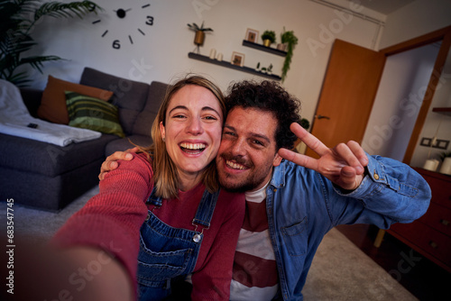 Cheerful couple at home using smartphone taking selfie to post on social media, having fun together in new apartment. POV people lifestyle. photo