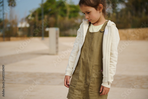 Portrait of a child girl in khaki dress and beige hoodie, looking down, expressing sadness standing on urban background © Taras Grebinets