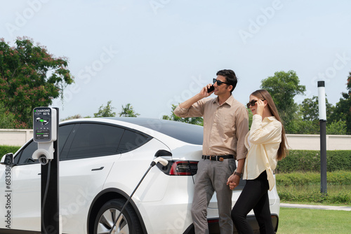 Young couple recharge her EV electric vehicle at green city park parking lot while talking on phone. Sustainable urban lifestyle for environment friendly EV car with battery charging station.Expedient © Summit Art Creations