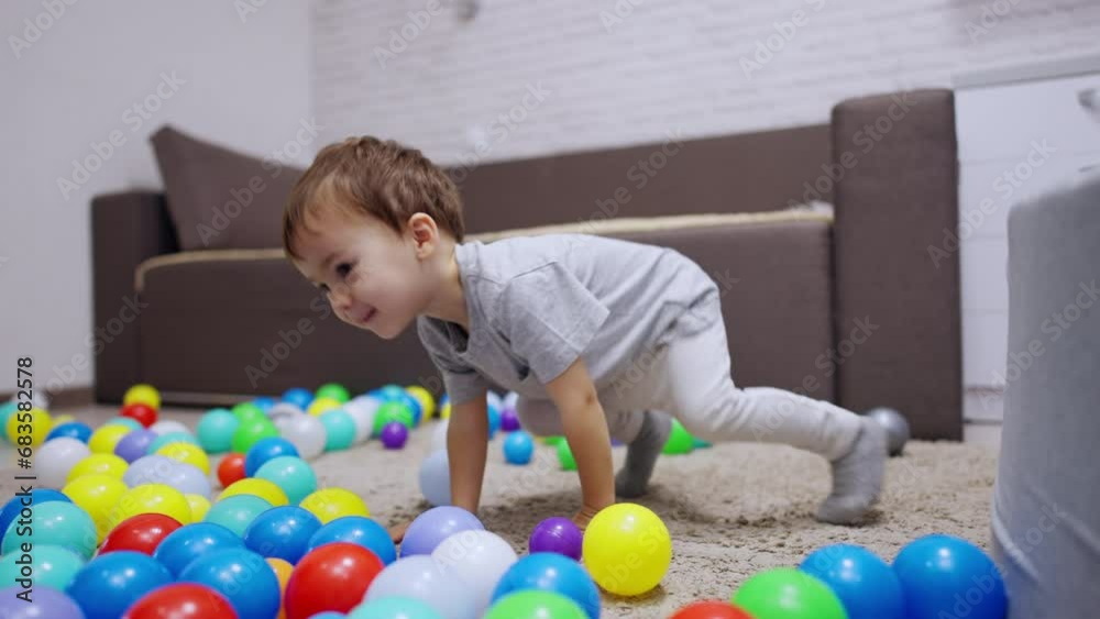 Kid in a grey t-shirt sits on the floor in his room. Lovely toddler jumps among the balls scattered by the carpet. Kid in a grey t-shirt sits on the floor in his room. Lovely toddler jumps among the balls scattered by the carpet.