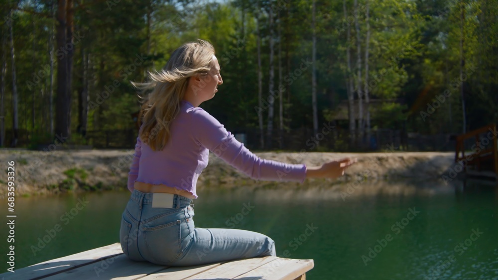 Young woman sits on pier and throws stone. Stock footage. Beautiful young woman sits on edge of pier by lake in summer. Woman on lake pier on sunny summer day