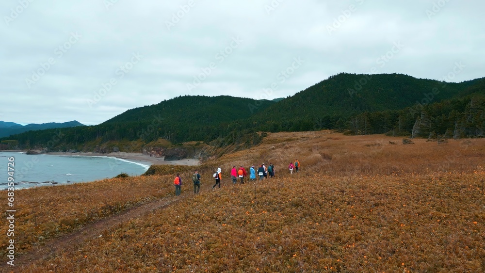 Top view of tourists walking along mountain coast trail. Clip. Hikers walk along mountain coast road on cloudy day. Tourists walk along northern mountain coast on background of sea and forests