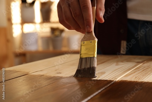 Man with brush applying wood stain onto wooden surface indoors, closeup photo