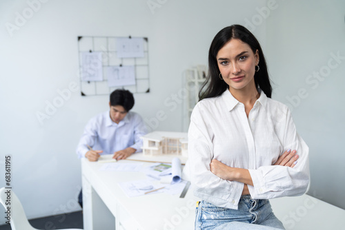 Closeup of female professional architect engineer poses with confident and looks at camera while skilled coworker focus on drawing blueprint. Creative living and design concept. Immaculate.