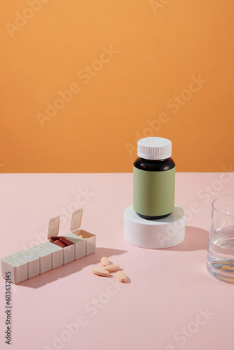 An unbranded medicine bottle is placed on a round podium, a daily pill box and a glass of water on a pastel pink table with an orange background. Drug advertising.