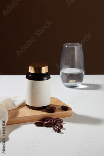 An unlabeled medicine bottle is placed on a wooden platform with soft capsules and a glass of water on a white table. Dark brown background. Mockup for dietary supplement advertising.