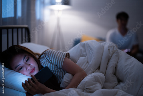 Asian girlfriend reading cartoon before watching Korean drama series while lie down and turn back in bed because sulk and angry boyfriend check online work on a sofa when bedtime night not go to sleep photo