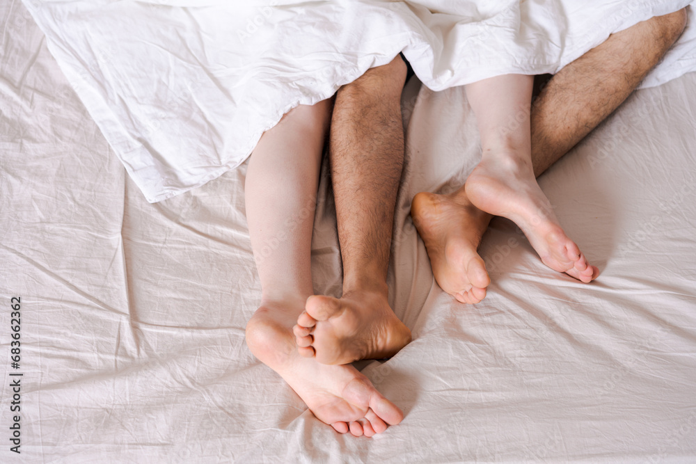 Feet of a couple in bed under the white blanket. - obrazy, fototapety, plakaty 