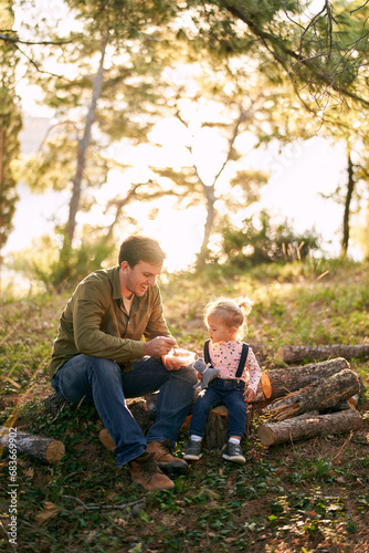 Little girl sits on a stump near her dad and watches as he picks up porridge from a lunchbox with a spoon © Nadtochiy