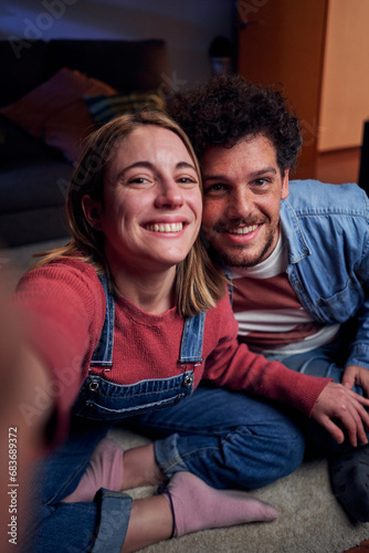 Cheerful couple at home using smartphone taking selfie looking at camera to post on social media, having fun together in new apartment. Vertical POV people lifestyle. © Gigi Delgado