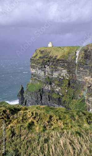O'Brien's Tower, which overlooks the Atlantic Ocean, is enhanced by the enormous water stream and an unexpected rainbow created as waves crash on the rugged Cliffs of Moher. Co. Clare, Ireland