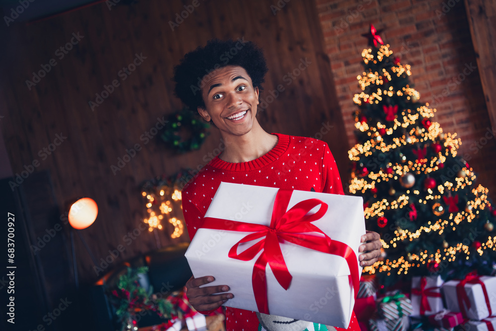 Photo of cheerful positive guy wear red ugly xmas sweater preparing christmas present indoors apartment room