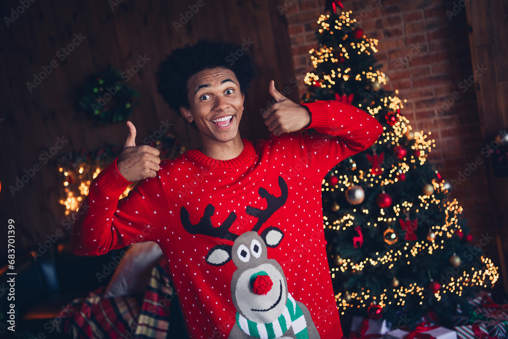 Photo of impressed excited guy wear red ugly xmas sweater showing two thumbs up recommend christmas party indoors apartment room