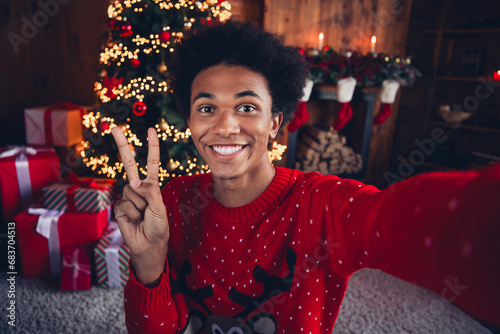 Photo of funky cheerful guy wear red ugly xmas sweater showing v-sign recording video christmas vlog indoors apartment room © deagreez