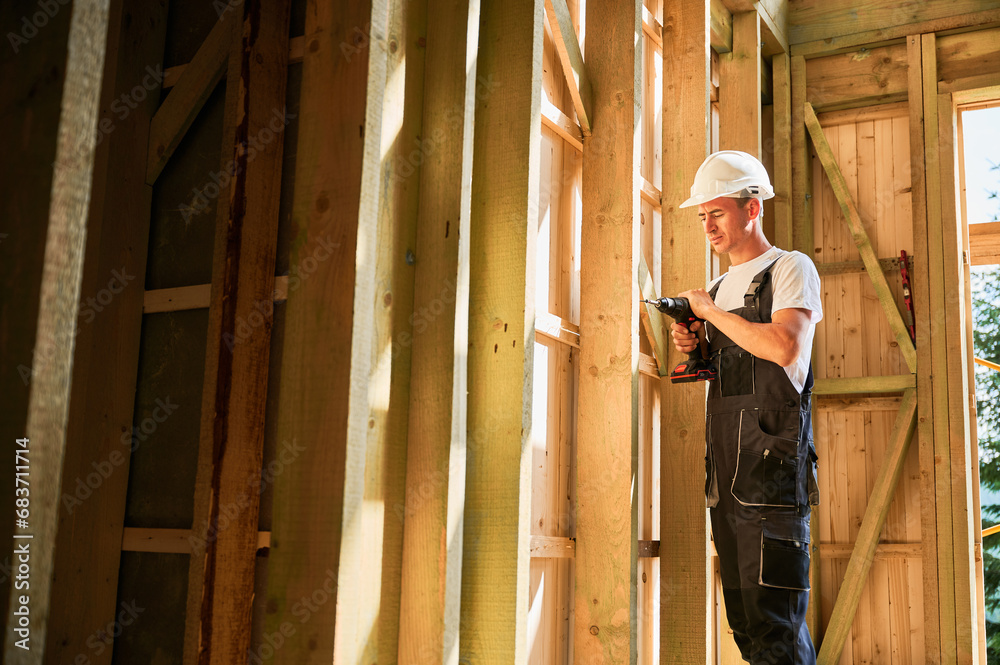 Carpenter constructing wooden framed house. Man worker working with screwdriver, wearing work overalls and helmet. Concept of modern eco-friendly construction. Carpenter constructing wooden framed house. Man worker working with screwdriver, wearing work overalls and helmet. Concept of modern eco-friendly construction.