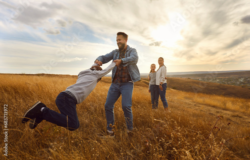Happy parents with two kids walking together at sunset and having fun in the field. Young smiling father playing with his boy child outdoors. Family walk in nature and leisure concept.