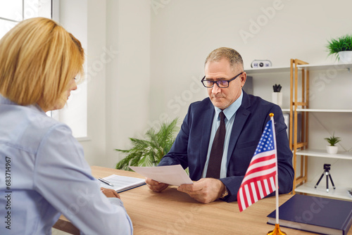 Mature male worker at USA consulate or visa agency sitting at office table with American flag, reading visa application of woman customer who wants to travel or immigrate to United States of America