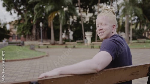 Homem albino, com cabelo dreadlocks, sorrindo, sentado no banco da praca em um dia ensolarado. Cinematico 4k. photo