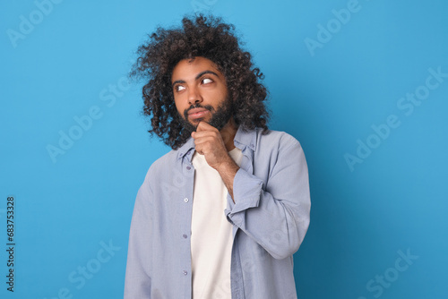 Valokuva Young thinking curly Arabian man scratches chin and thinks about plan for next week or is trying to come up with idea for launching own money-making business stands posing in blue studio