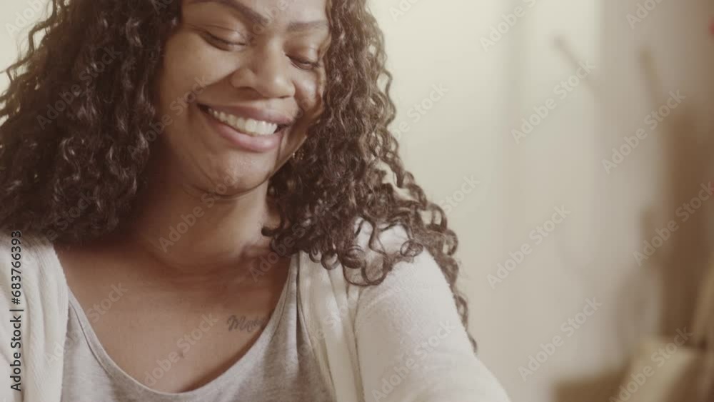 Mulher sorridente com cabelos cacheados, sentada na cama, aplicando hidratante na pele. Cinematico 4k