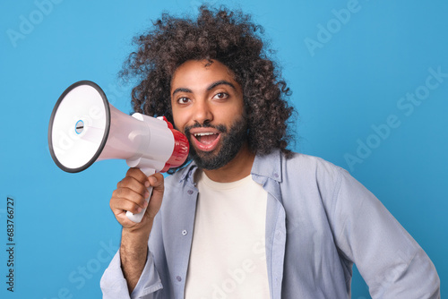 Young smiling energetic Arabian man promoter with megaphone announces start of sales of new product or calls to become reader of news site dressed in casual clothes stands on blue background.