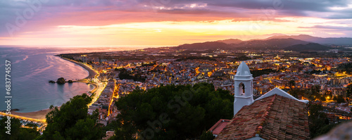 Panoramic photo from Sant Joan Hermitage, Blanes, with its white walls in the foreground and the illuminated city under a vibrant twilight sky.