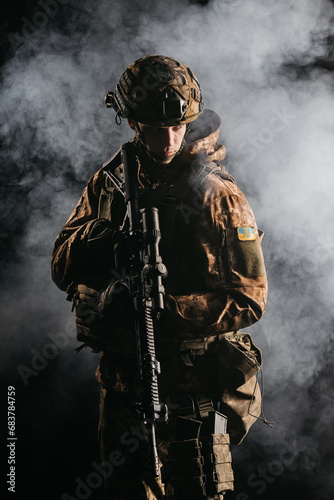 An army soldier in a military camouflage uniform, a helmet and a mask, holds a rifle and aims with a red dot sight, standing in an attack position, in the smoke on a black background. © Vladislav
