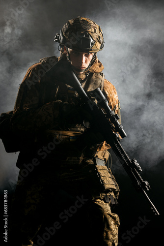 An army soldier in a military camouflage uniform, a helmet and a mask, holds a rifle and aims with a red dot sight, standing in an attack position, in the smoke on a black background.