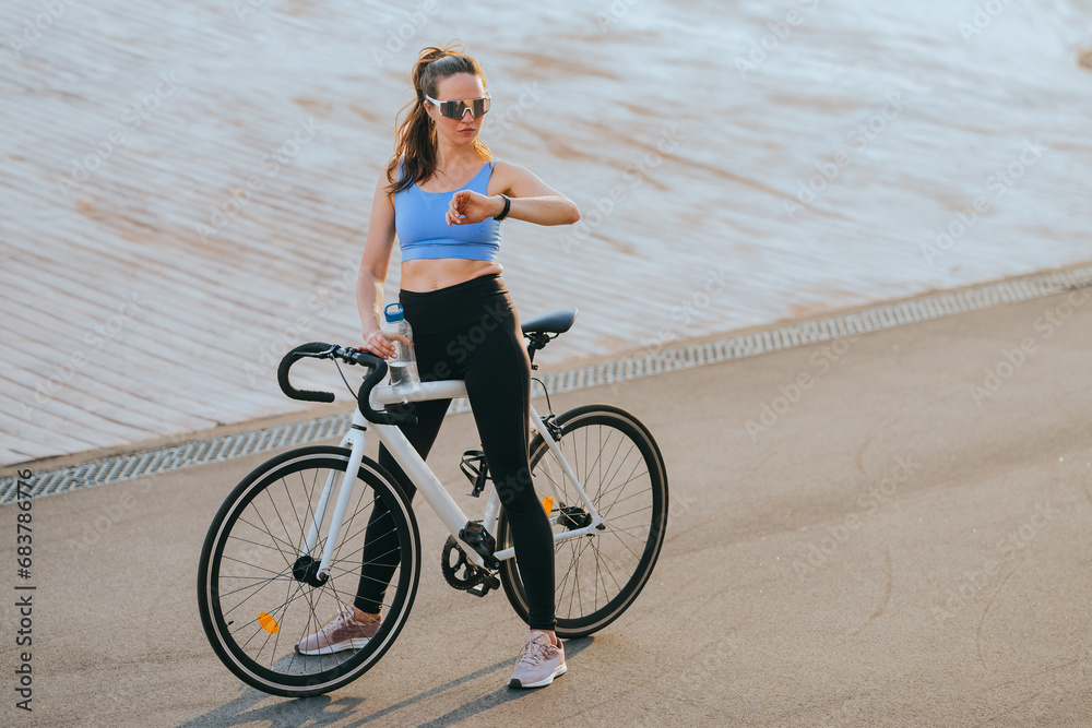 Fototapeta premium A beautiful young American girl in sports leggings, blouses in sunglasses is sitting on a bicycle, will make a stop in training, look at a fitness bracelet, check her pulse.